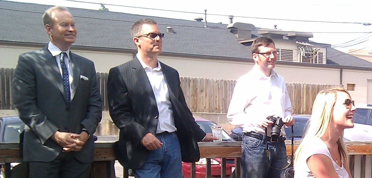 A group of light skinned people gathered on the outdoor deck of a cafe, some seated at tables while others lean on the railing. They're talking and smiling.