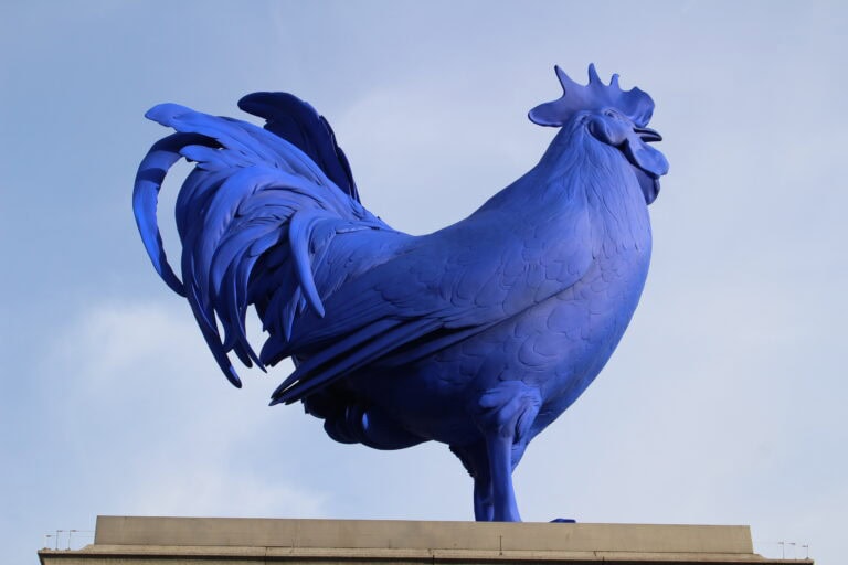 A large bright blue rooster sculpture mounted on a stone plinth against a pale blue sky, photographed in Trafalgar Square, London in 2013.