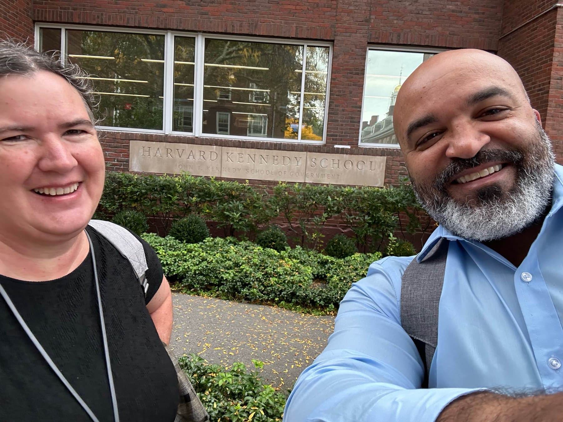 A light skinned woman and a medium skinned man with a gray beard stop for selfie in front of a brick sign that says Harvard Kennedy School