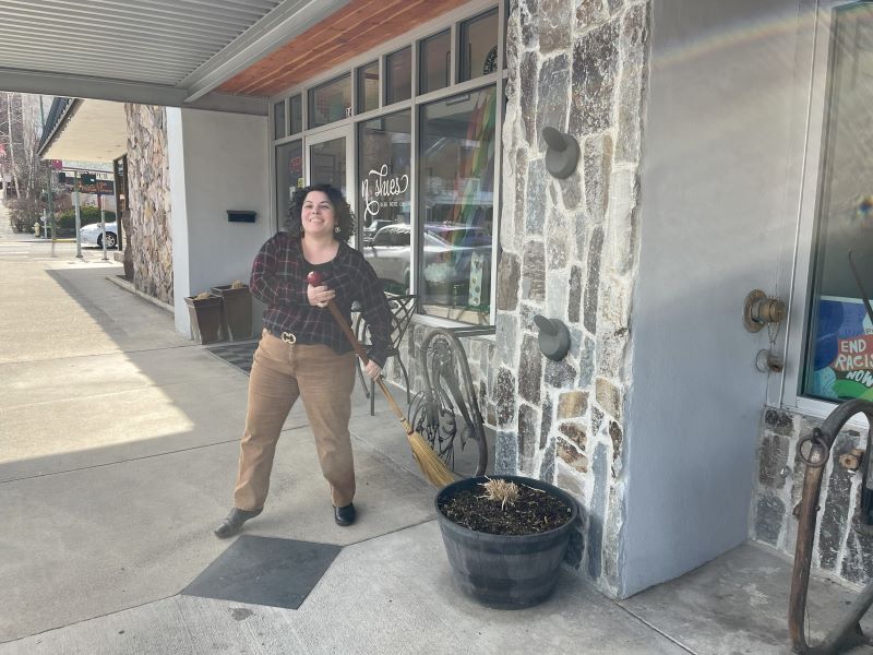 A woman with dark curly hair is sweeping the sidewalk in front of her downtown businesses.