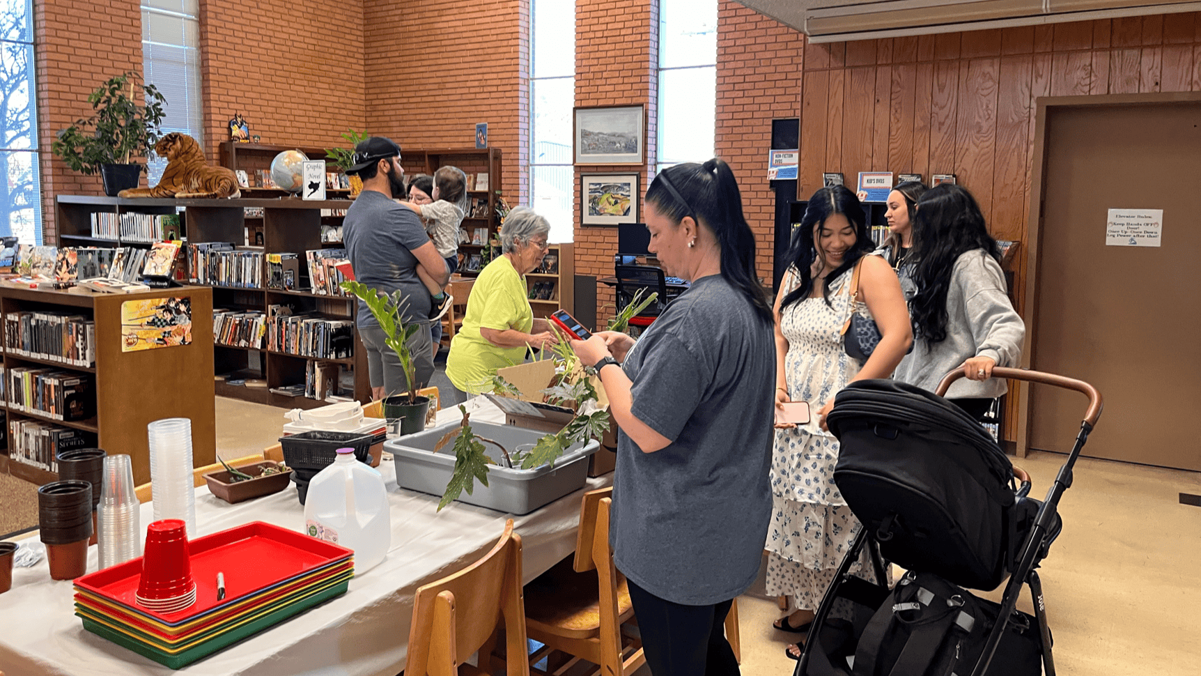 Diverse group gathers at library plant swap. Woman in foreground examines potted plant while people of various ages browse plants on tables. Baby stroller visible. Bookshelves and brick walls frame the library space. Colorful trays stacked on table.