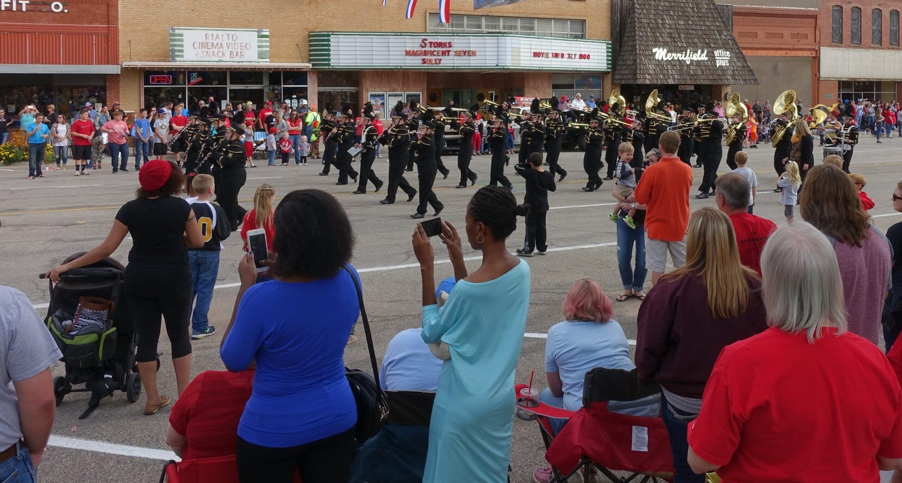 A diverse group of people line the street in a small town, watching a marching band in a parade.