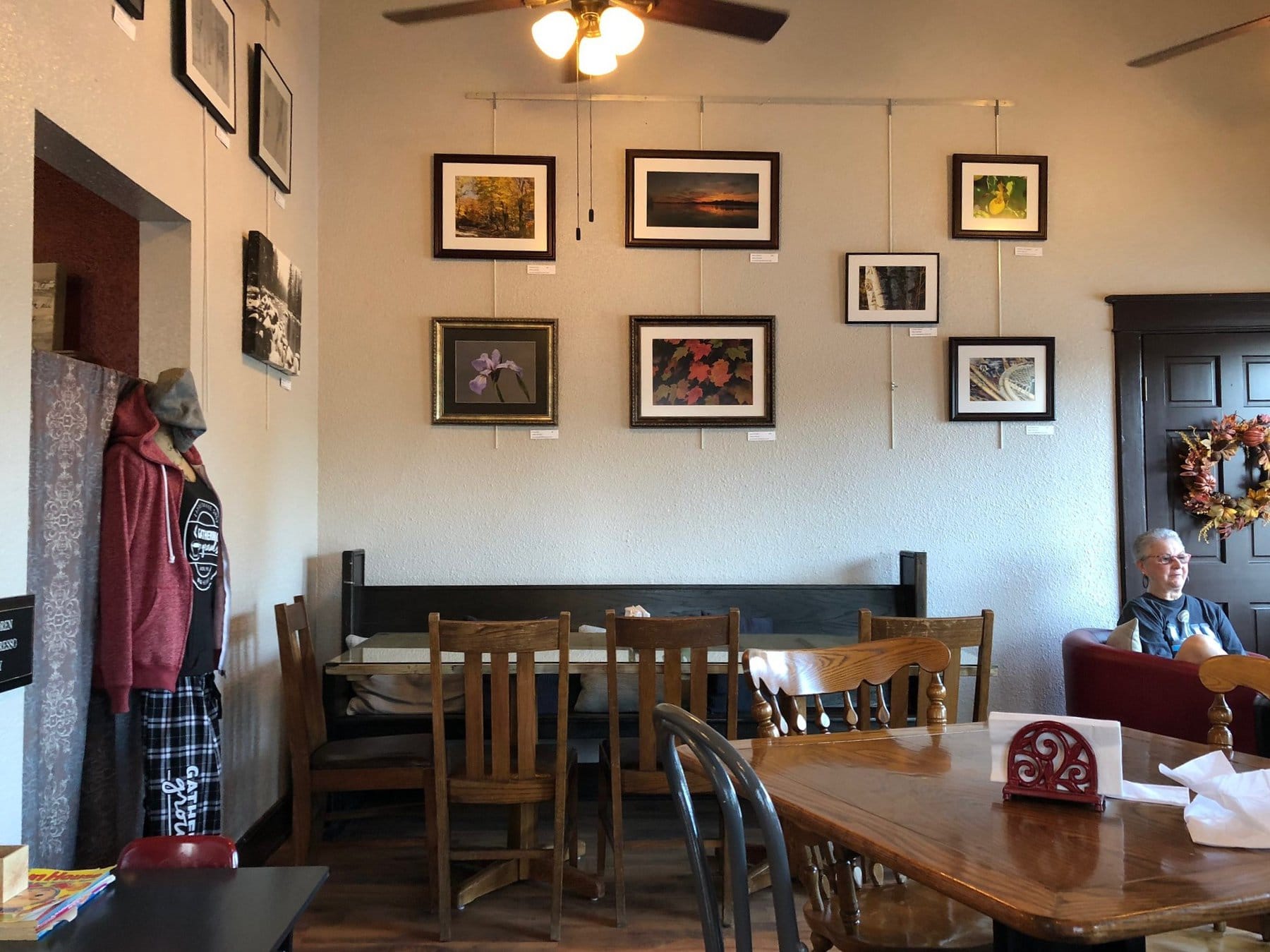 Local art is nicely framed and hung gallery style on the walls of a local coffee shop. A white-haired woman is sitting at one table.