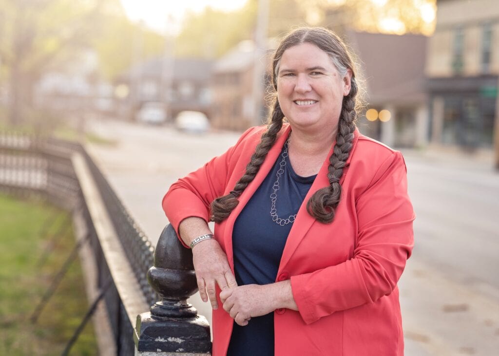 Becky McCray smiles warmly with long braided hair and coral blazer. A row of small town storefronts blur in the background.