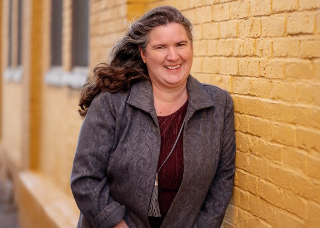 Becky McCray smiling, standing next to a yellow-painted brick wall