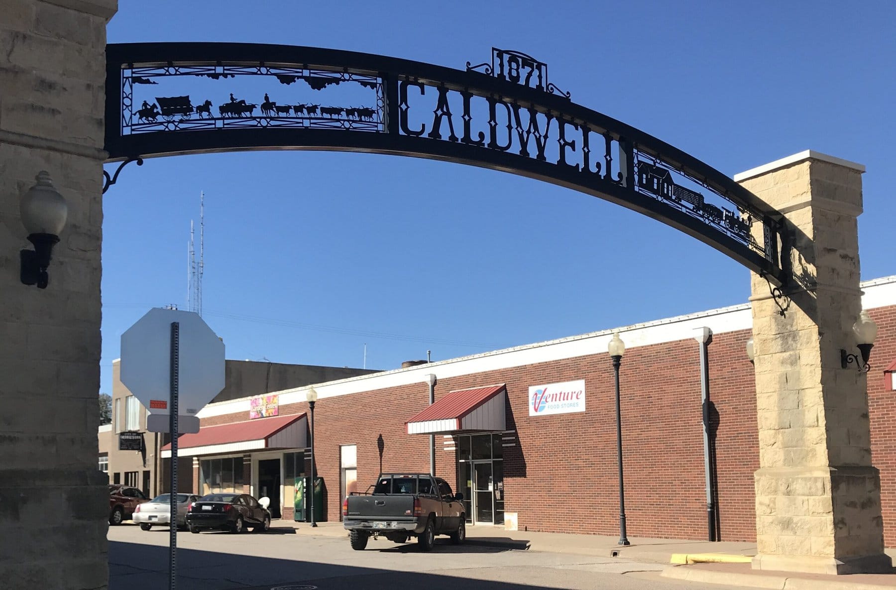 A metal archway over a street is decorated with a cattle drive on one side and railway loading corral on the other. The center says "Caldwell, 1871". The arch rests on two massive limestone pillars. Downtown buildings are visible in the background.