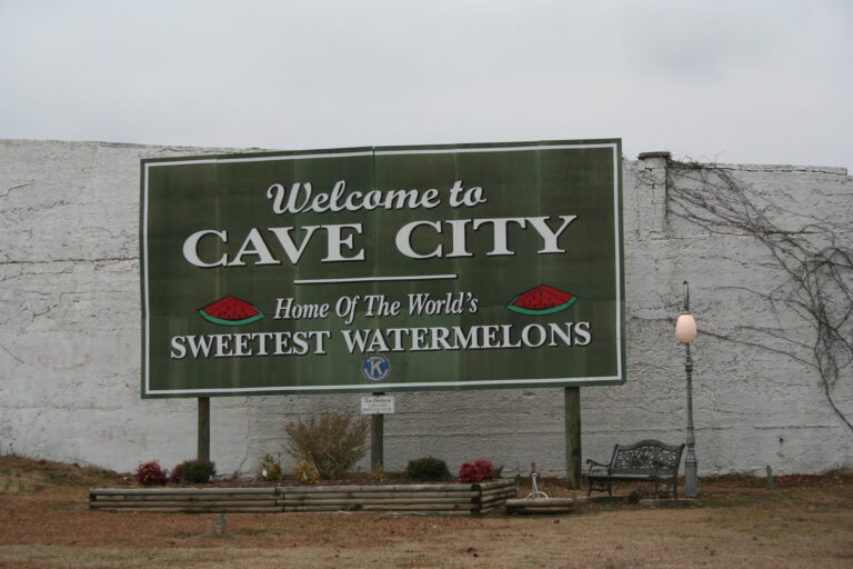 A green billboard reading "Welcome to Cave City, Home of the World's Sweetest Watermelons" with watermelon slice graphics, mounted in front of a whitewashed wall with a park bench and lamppost nearby.