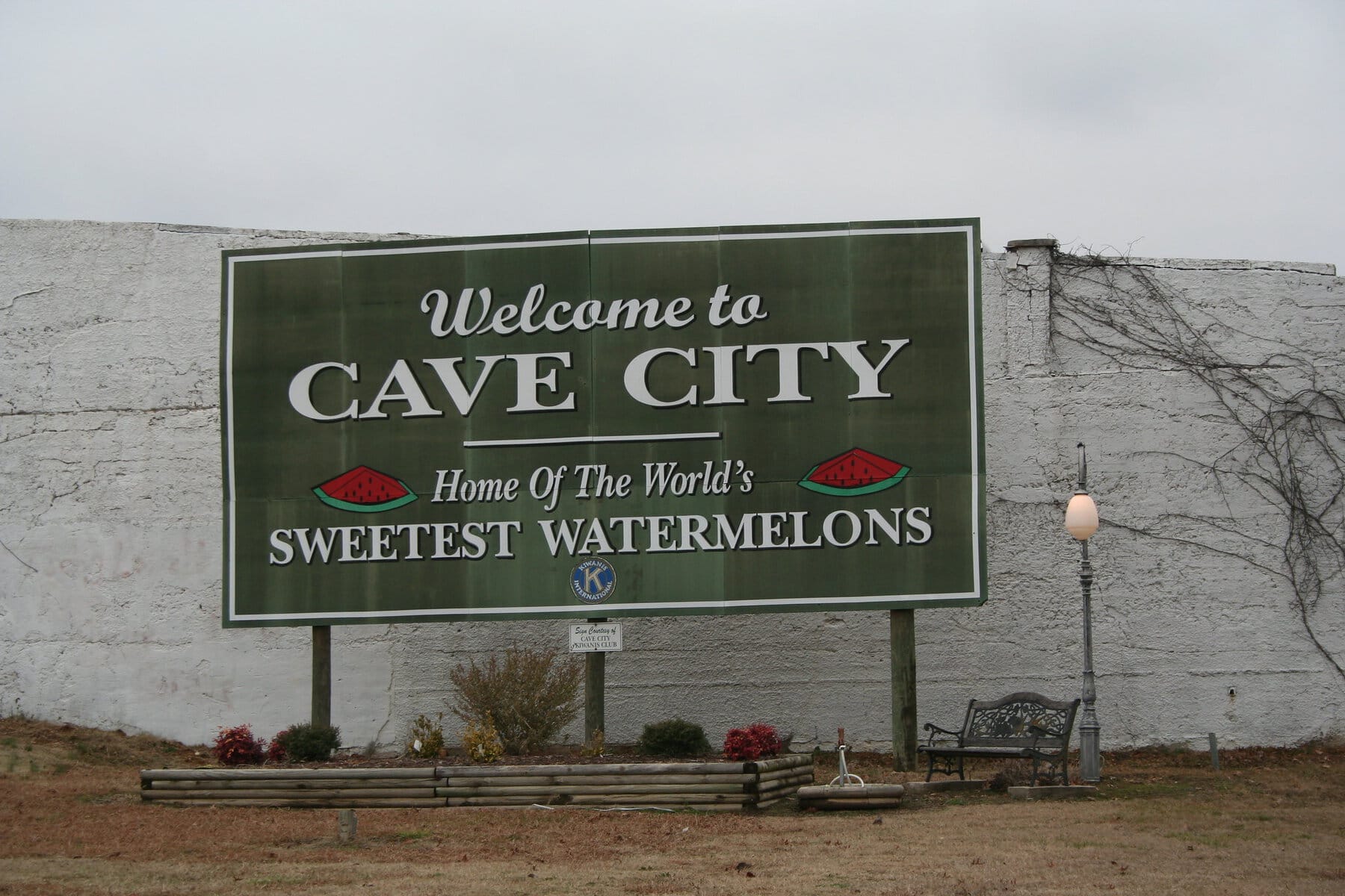 A green billboard reading "Welcome to Cave City, Home of the World's Sweetest Watermelons" with watermelon slice graphics, mounted in front of a whitewashed wall with a park bench and lamppost nearby.