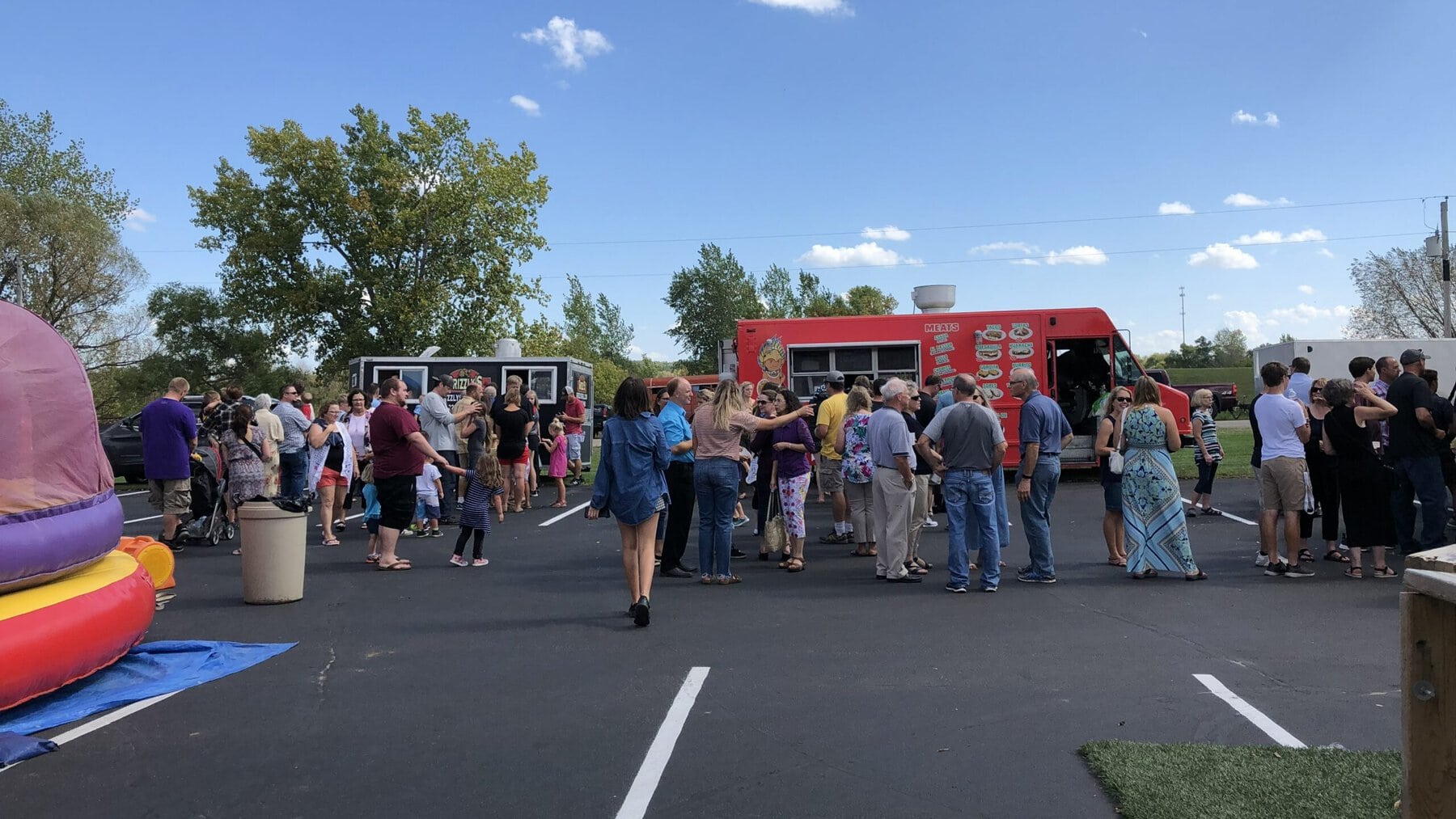 A large crowd of people of all ages gathered around food trucks in a parking lot at a rural community event, with a bouncy house on the left and trees and open sky in the background.