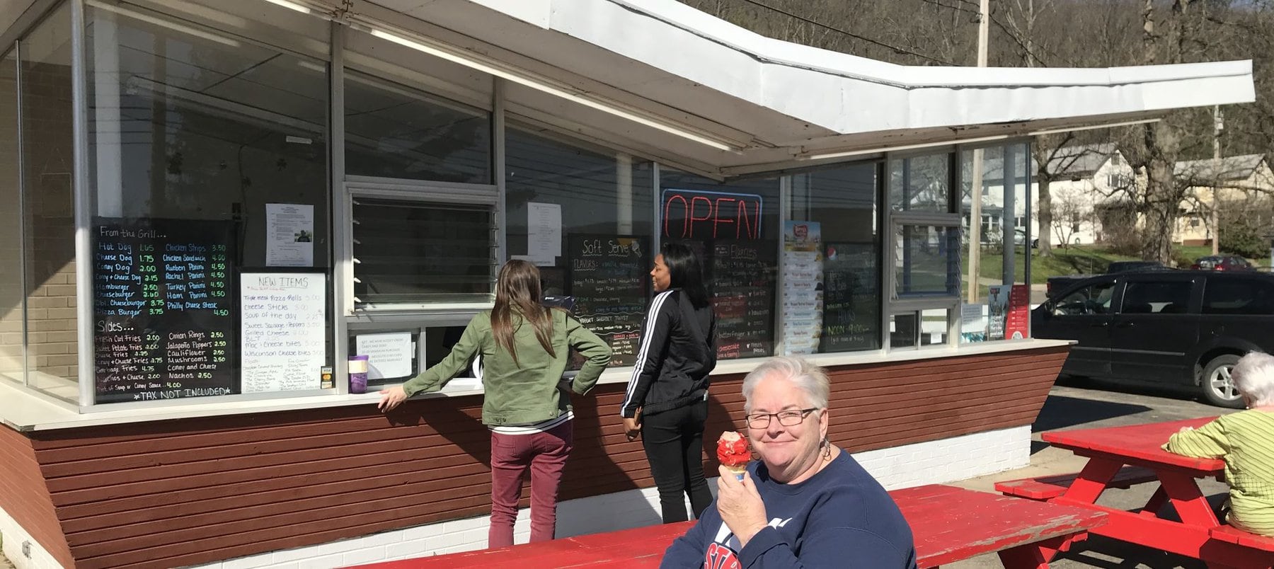 Old fashioned 1950s style walk up ice cream place with picnic tables out front and a diverse group of women customers. An older woman enjoys an ice cream cone covered in cherry red dip. Two younger women are waiting for their order. Another woman is seated to one side.