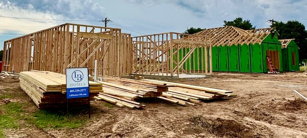 Two houses are shown in the framing stage. In the foreground, one house has the stud walls up. A stack of lumber and a real estate sight sit in front of it. In the background, another house has rafters up and the side walls are wrapped in greenboard.