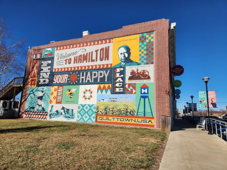 Large colorful mural on brick building reads "Welcome to Hamilton" and "Your Happy Place." Quilt patterns, local imagery including JC Penney portrait, steam engine, livestock, corn, and water tower depicting "Quilttown USA." Matching banners visible on streetlights along wide sidewalk.