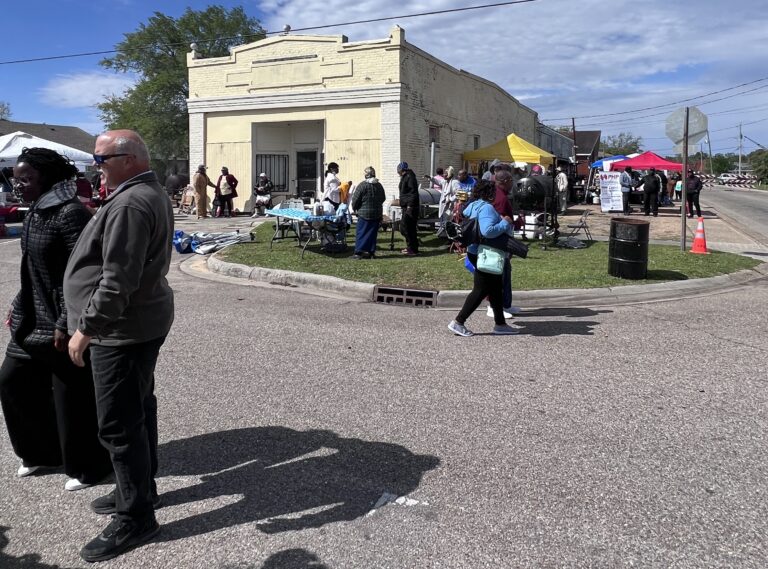 A street fair or market is happening near a single downtown-style building. People are shopping at booths and others are walking or holding conversations in the street. Most are Black or dark skinned.