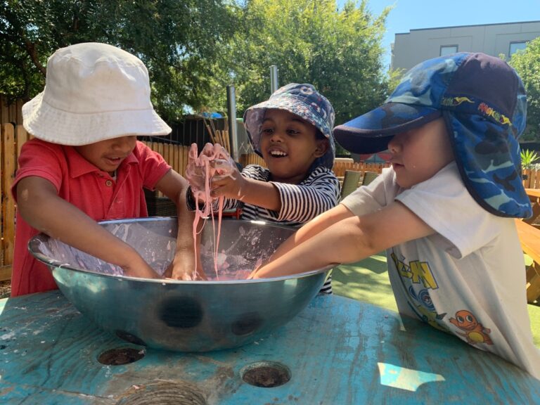 Three diverse young children in sun hats laugh and play with pink oobleck in a metal bowl outdoors. The child in the center lifts dripping oobleck with both hands.