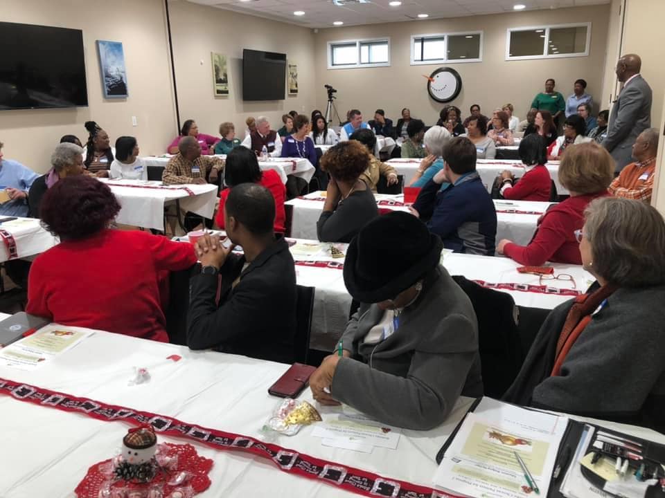 A diverse group of people seated at long tables are joining in conversation and making notes.