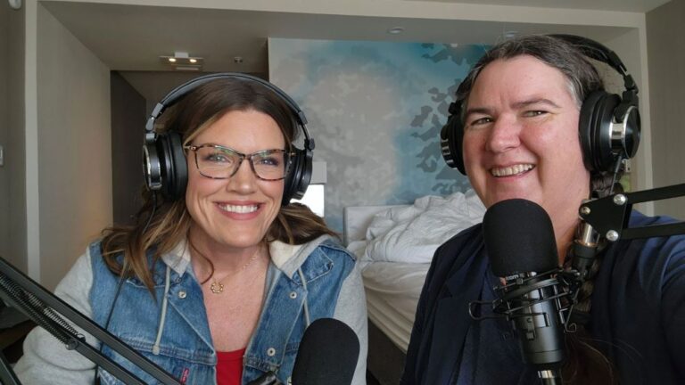 Two light-skinned women with dark hair wearing headphones with microphones in front of them on boom arms. Both are smiling and looking at the camera.