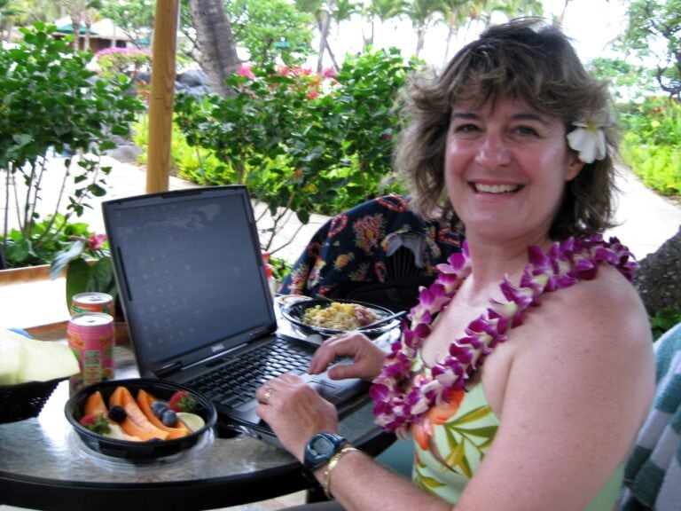 A happy, smiling light-skinned woman with light brown hair is wearing a bathing suit with a lei and has a flower tucked behind her ear. She's seated at a shady poolside table with a laptop open in front of her, along with a dish of fruit and a few beverages.