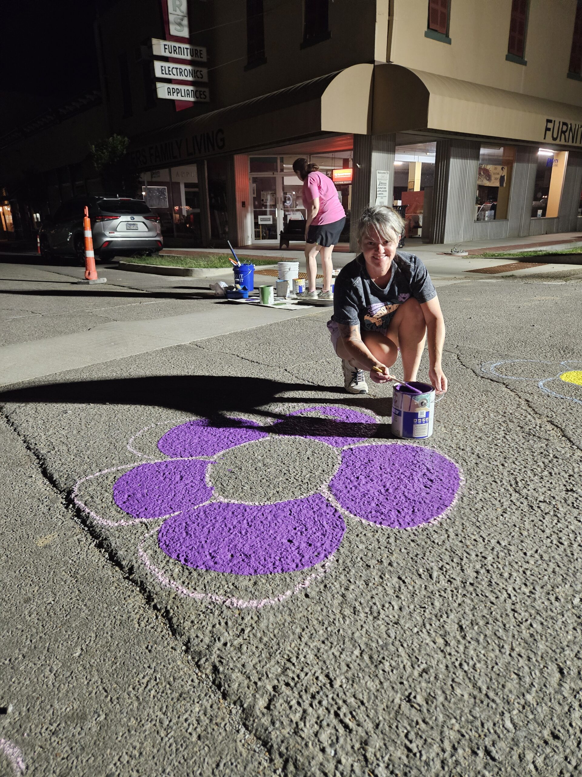 Two women are painting flower designs on the asphalt in an intersection. The sky is still dark, and they are lit by bright worklights.
