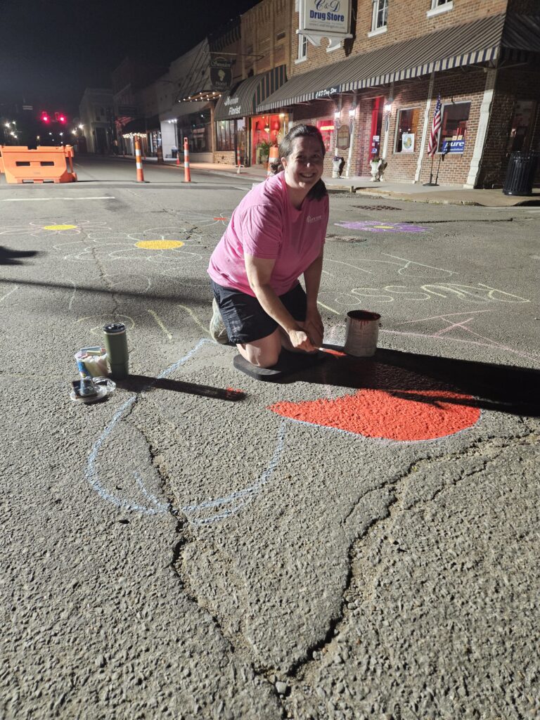 A woman smiles while she paints a red heart on the asphalt in an intersection. The sky is still dark, and she is lit by bright worklights.