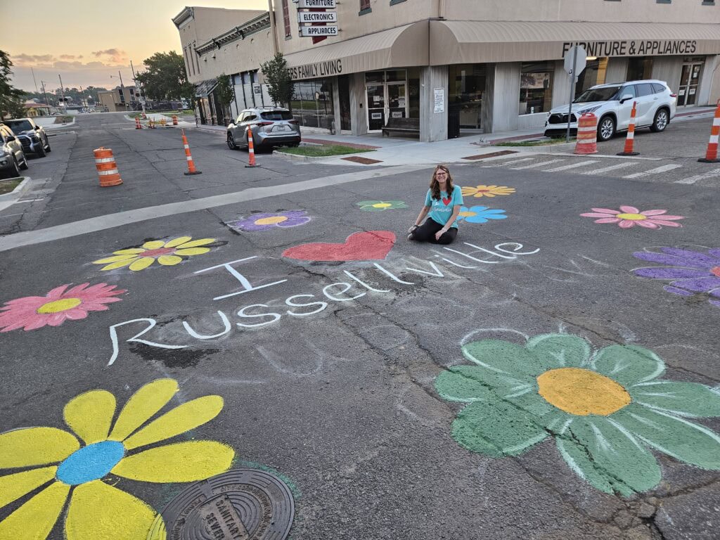 Sara Jondahl is sitting in the middle of the painted intersection at dawn, surrounded by painted flowers and the message I heart Russellville.