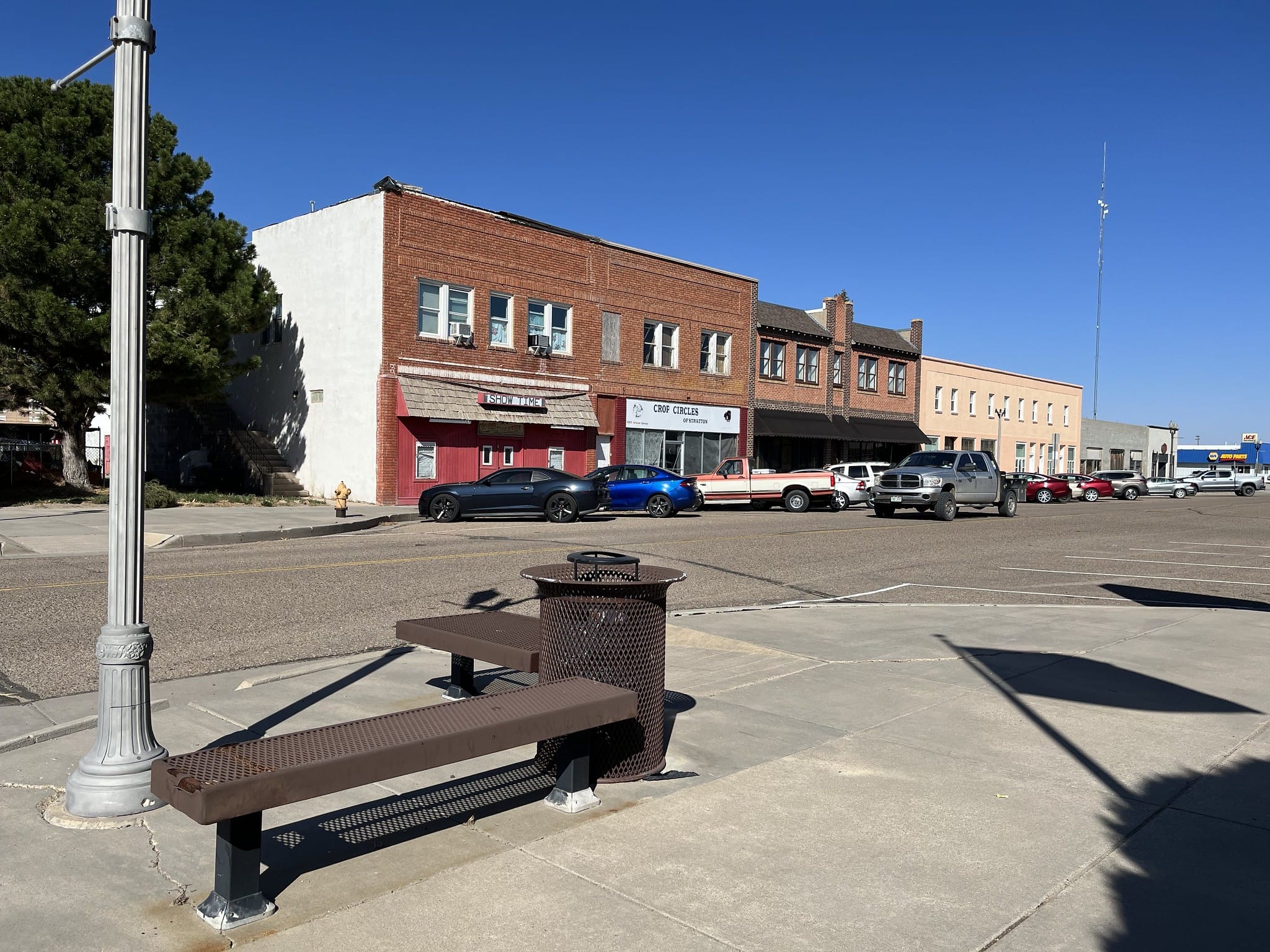 A small town downtown with brick-fronted buildings. On the corners are benches and old fashioned light poles.