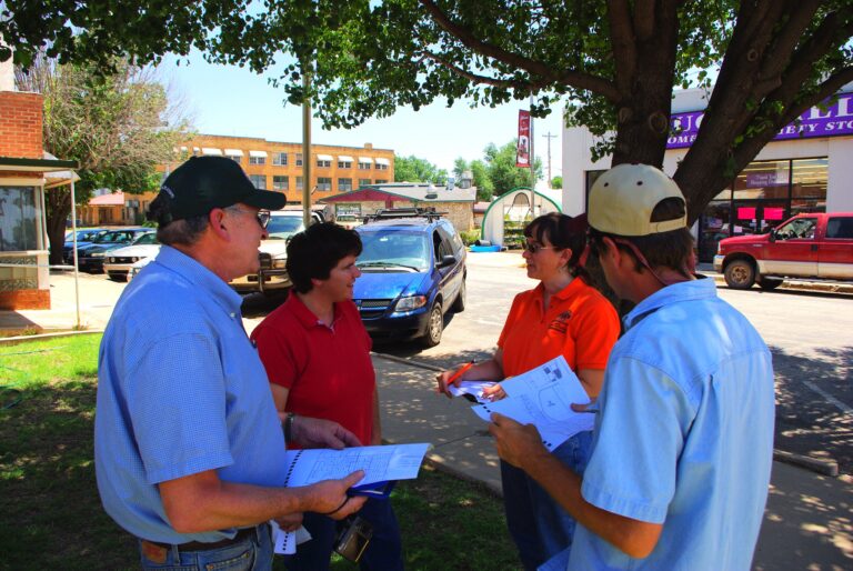 Four people standing together under a shade tree on a small town main street, reviewing planning documents.