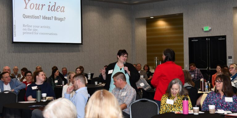 A woman holding a microphone sharing an idea with a full room of community foundation leaders, while Becky McCray in a red jacket listens nearby and a SaveYour.Town slide reading "Your idea — Question? Ideas? Brags?" is projected on screen behind them.