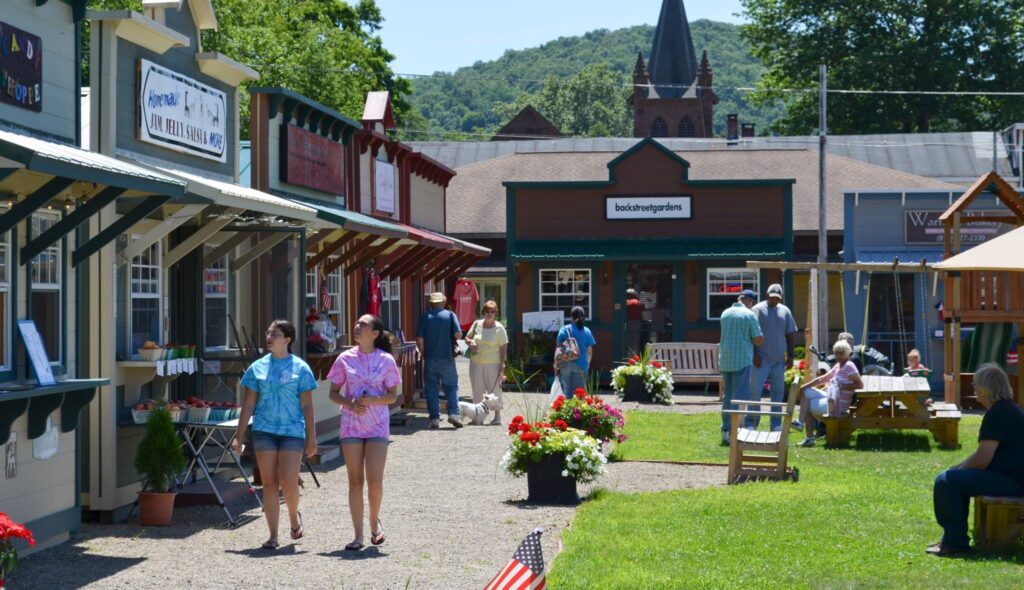 A row of brightly painted sheds housing pop-up businesses. People are strolling in front of the businesses, while others gather and talk in the greenspace out front.