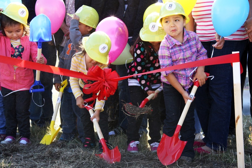 A group of pre-school age children wearing hardhats and holding shovels turn the first shovelfuls of dirt at a ceremony to celebrate the start of construction on a new Head Start Center on the Spirit Lake Reservation in North Dakota. Colorful balloons and ribbons surround the children.