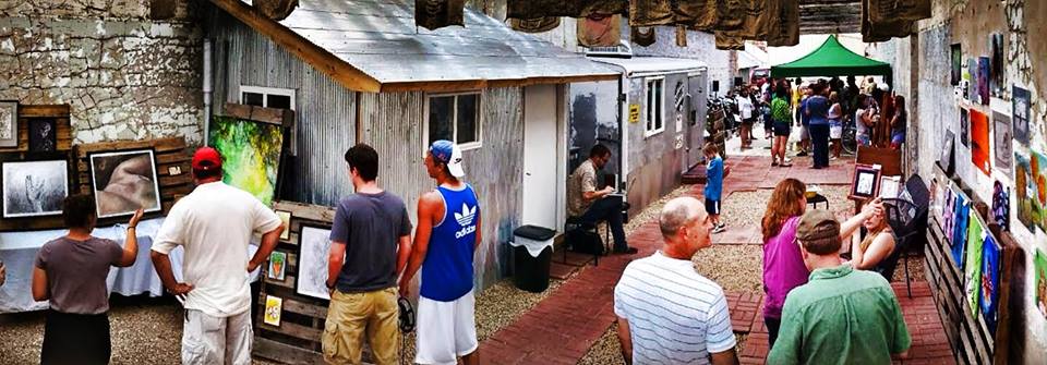 Crowd of people browsing an outdoor art exhibit inside a roofless brick building with corrugated metal walls, coffee sacks draped over exposed rafters