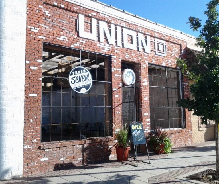 Exterior of the Union Square brick building in St. Francis, Kansas, with a Fresh Seven Coffee sign in the window, an open sign and potted plants out front, no glass in the window frames and no visible roofline behind the facade.