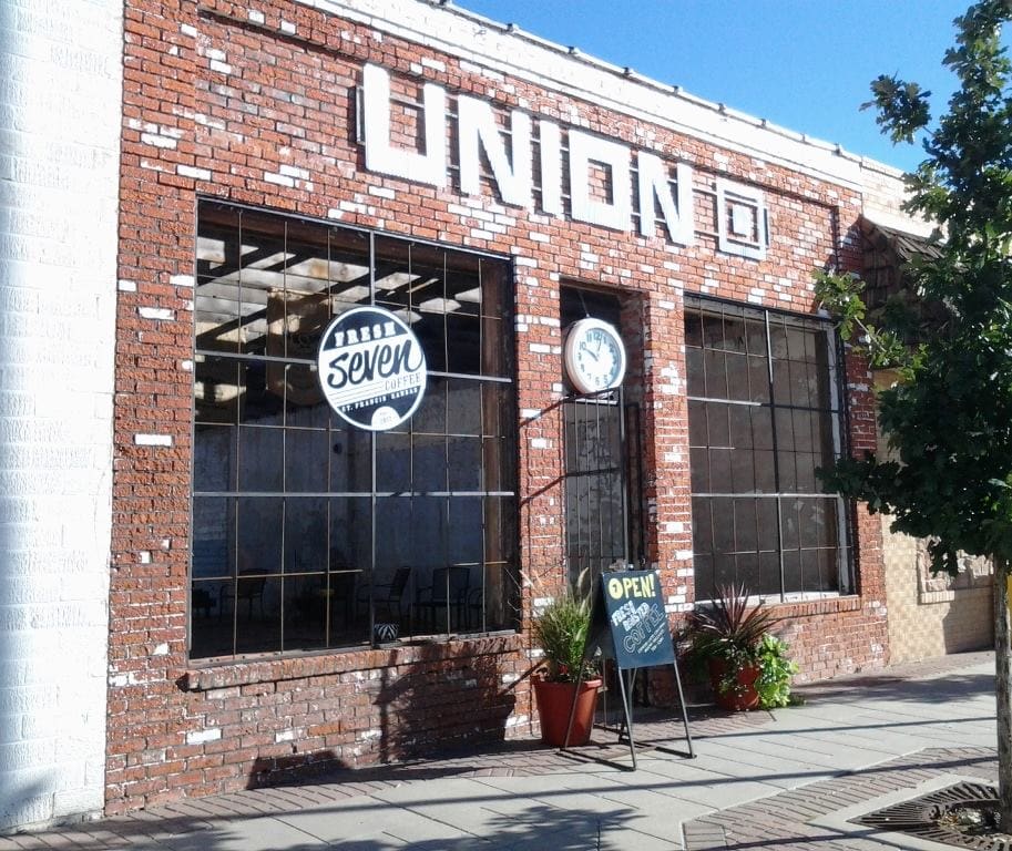 Exterior of the Union Square brick building in St. Francis, Kansas, with a Fresh Seven Coffee sign in the window, an open sign and potted plants out front, no glass in the window frames and no visible roofline behind the facade.