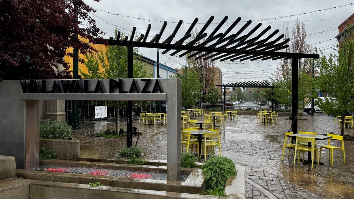 Plaza sign and water feature, with yellow tables and chairs on stamped concrete and a pergola with string lights overhead.