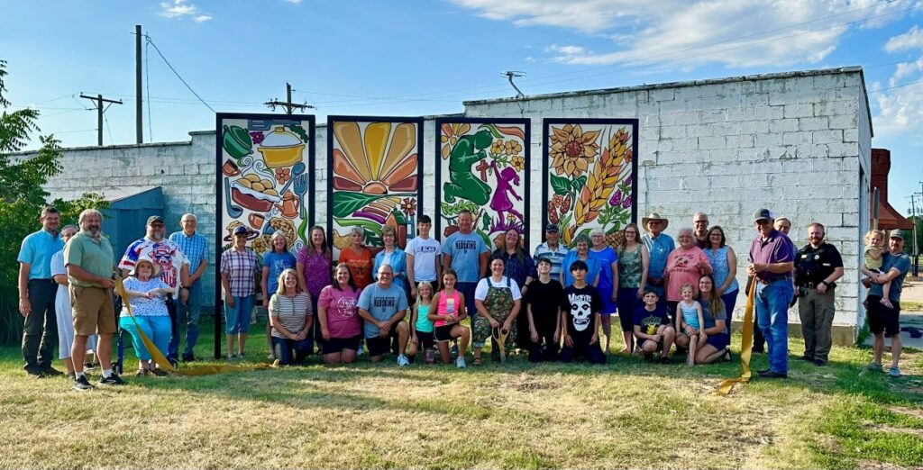 A large group of about 50 community members of all ages gathered in front of four colorful installed mural panels along a Main Street building in Windom, Kansas, holding yellow ribbon at the unveiling ceremony.