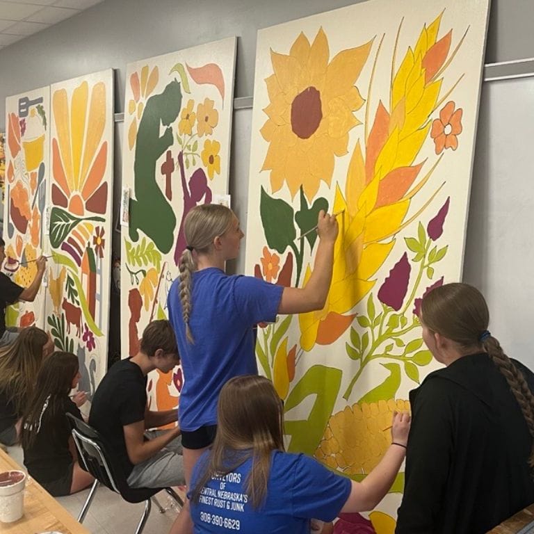 Several students in a classroom painting large colorful mural panels featuring food, agriculture and small town life in warm oranges, yellows and greens.