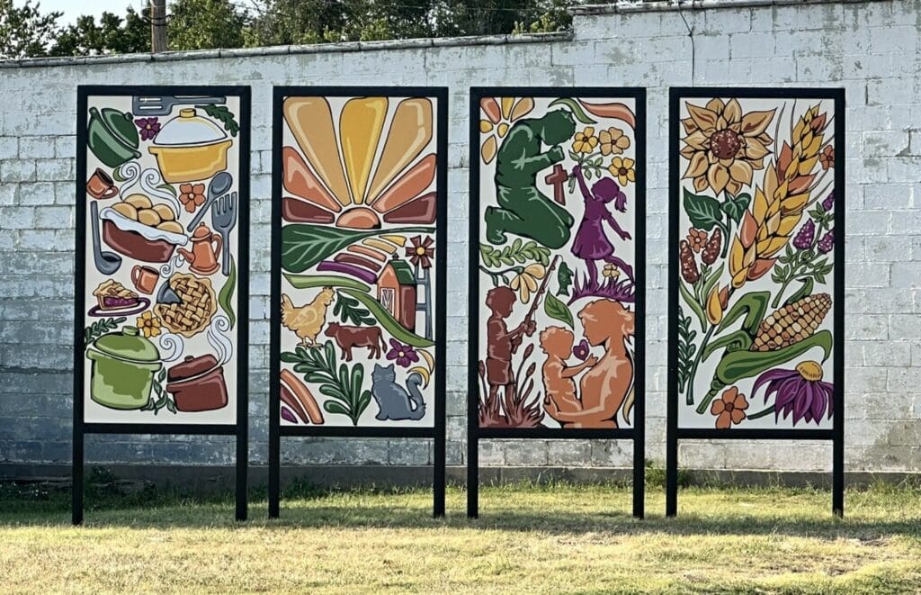 Four large freestanding colorful mural panels installed in front of a white painted building in downtown Windom, Kansas, depicting covered dish food traditions, farm life, community relationships and faith.