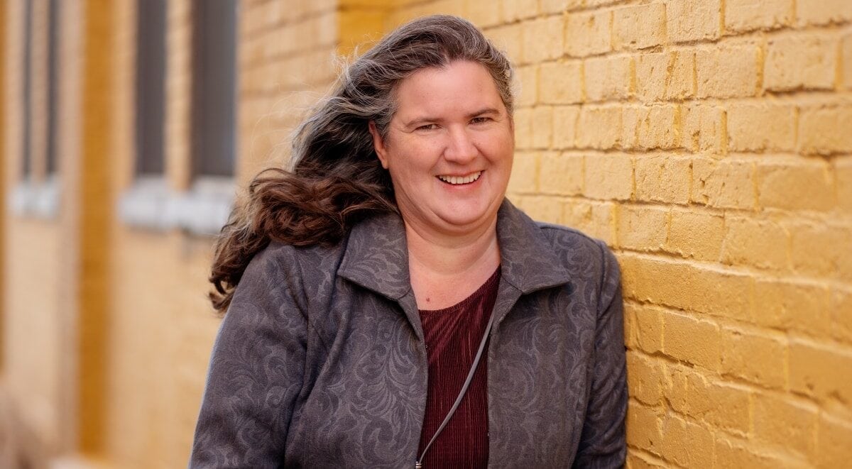 Becky McCray smiling, standing next to a yellow-painted brick wall