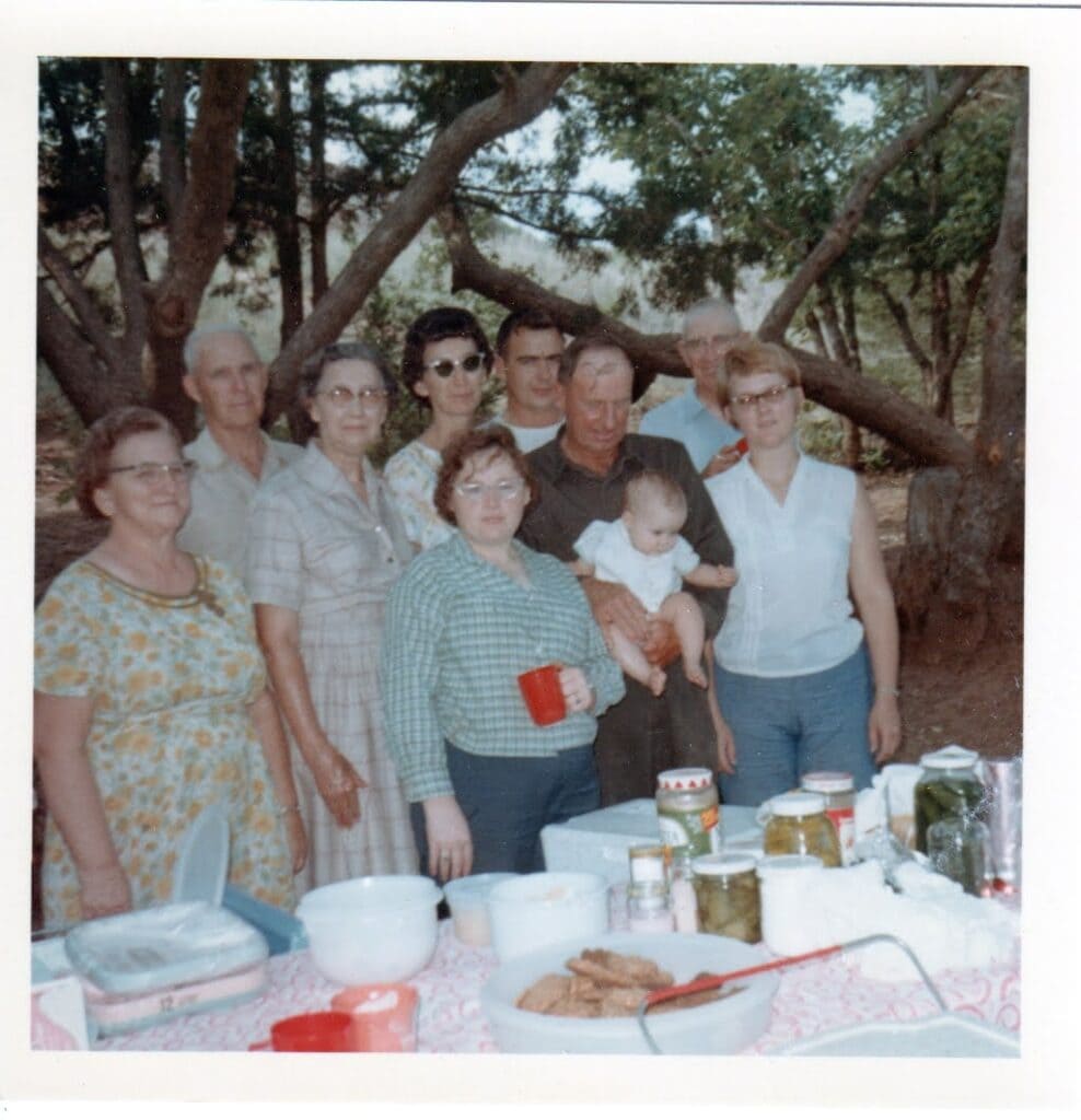 A 1960s multigenerational group, standing near a picnic table covered with dishes, home canned pickles and a big tupperware bowl full of cookies.