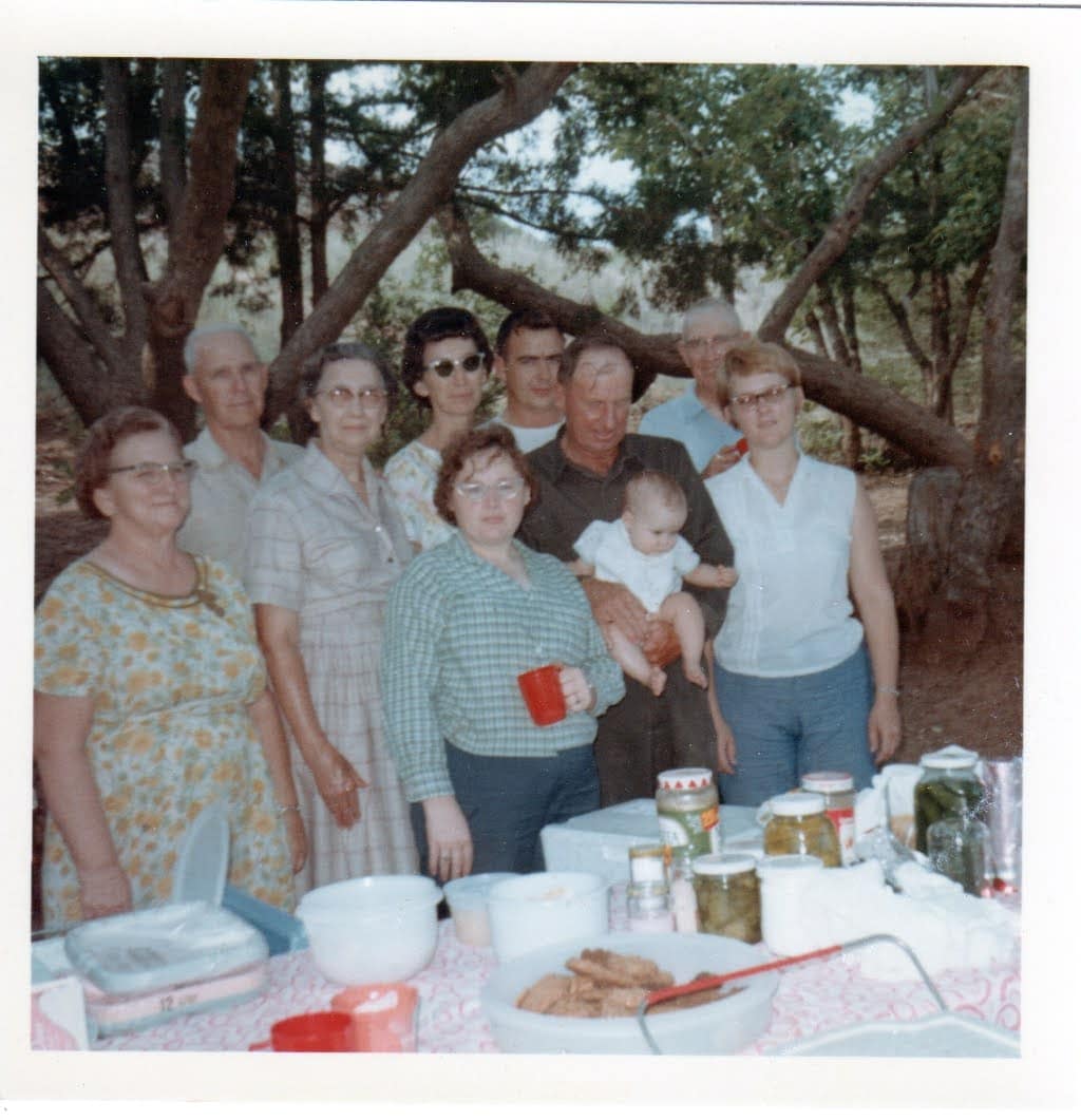 A 1960s multigenerational group, standing near a picnic table covered with dishes, home canned pickles and a big tupperware bowl full of cookies.