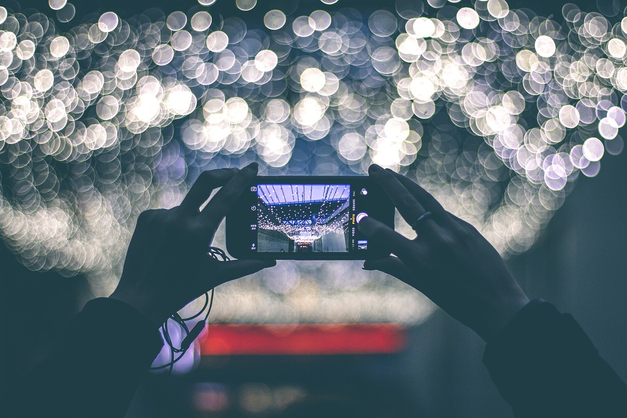 Hands holding a smartphone as a camera in the foreground. In the background string lights are shown in pleasing unfocused bokeh.